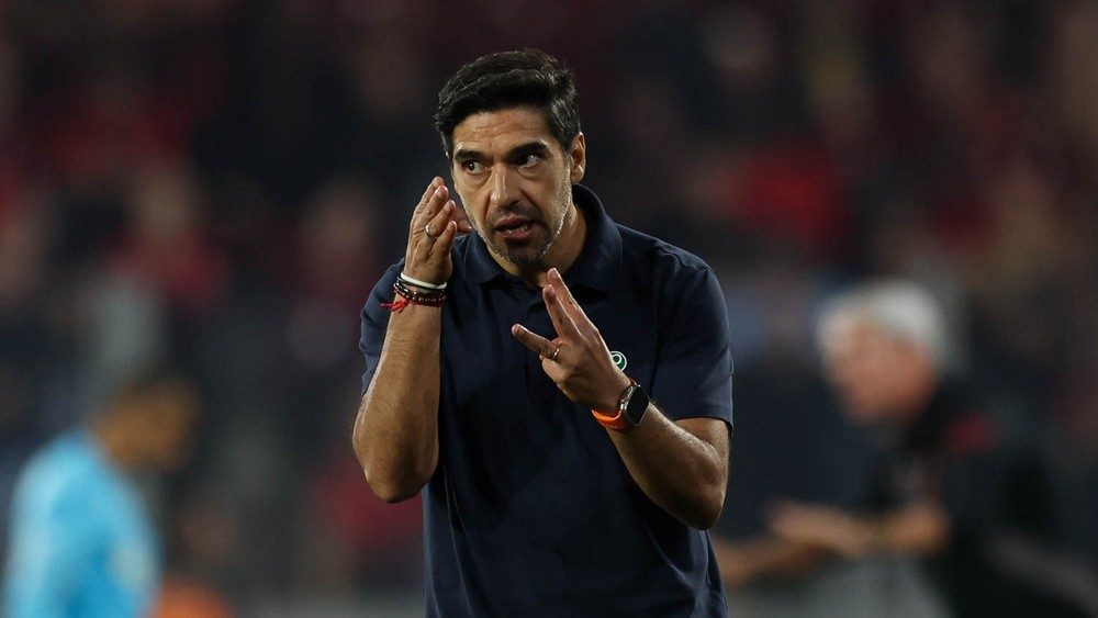 Abel Ferreira, de camisa polo azul-marinho com o escudo do Palmeiras, gesticula com as duas mãos à beira do campo, com a arquibancada desfocada ao fundo