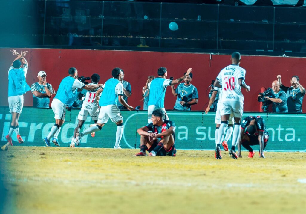 Jogadores do Vasco comemoram gol junto à beira do campo no Barradão, enquanto atletas do Vitória lamentam no gramado