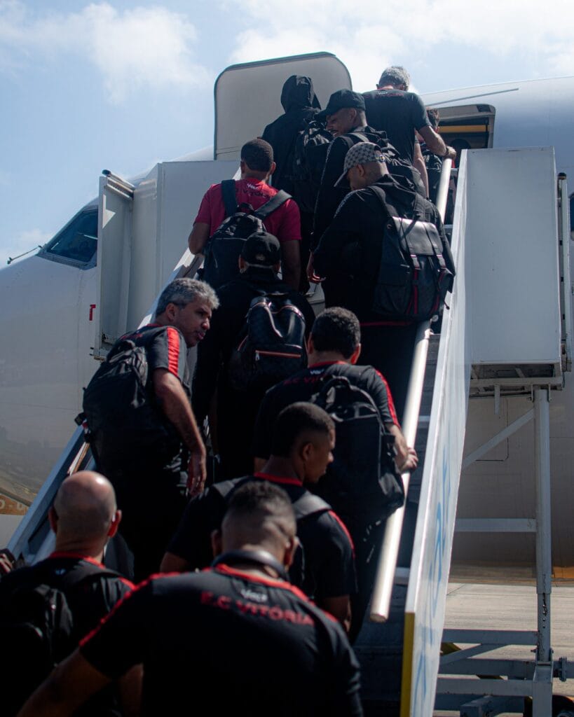 Jogadores e comissão do Vitória, de uniforme preto e vermelho e mochilas nas costas, sobem a escada do avião em viagem para jogo fora de casa