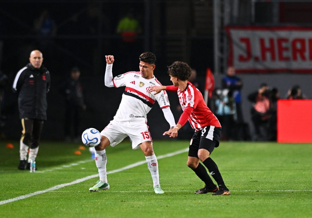 Jorge Carrascal, de camisa branca do Flamengo com o número 15, protege a bola sob marcação de um jogador do Estudiantes, de camisa listrada em vermelho e branco, no gramado do Estádio UNO