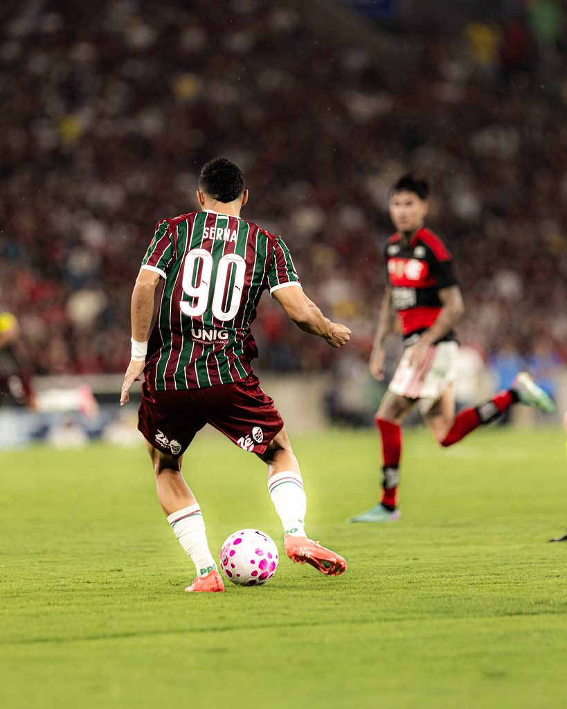 Kevin Serna, camisa 90 do Fluminense, conduz a bola de costas para a câmera no Maracanã, com um jogador do Flamengo ao fundo