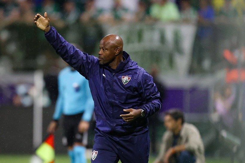 Marcão, técnico do Fluminense, de agasalho azul-marinho do clube, estende o braço direito e orienta o time à beira do gramado do Nubank Parque, com a torcida desfocada ao fundo