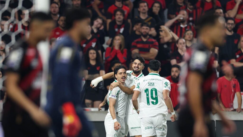 Maurício, de faixa na cabeça e camisa branca do Palmeiras, comemora o gol abraçado por um companheiro, com Sosa, camisa 19, de costas; jogadores do Athletico desfocados em primeiro plano e torcida rubro-negra ao fundo