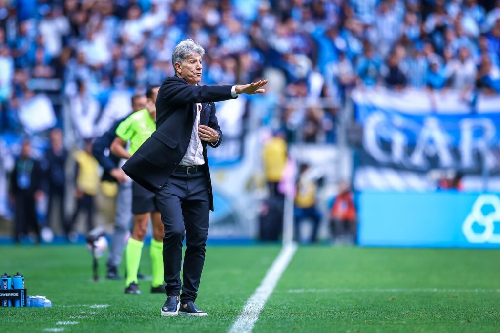 Renato Portaluppi, técnico do Grêmio, de terno escuro e camisa branca, estende o braço direito à beira do gramado da Arena do Grêmio, com a torcida ao fundo