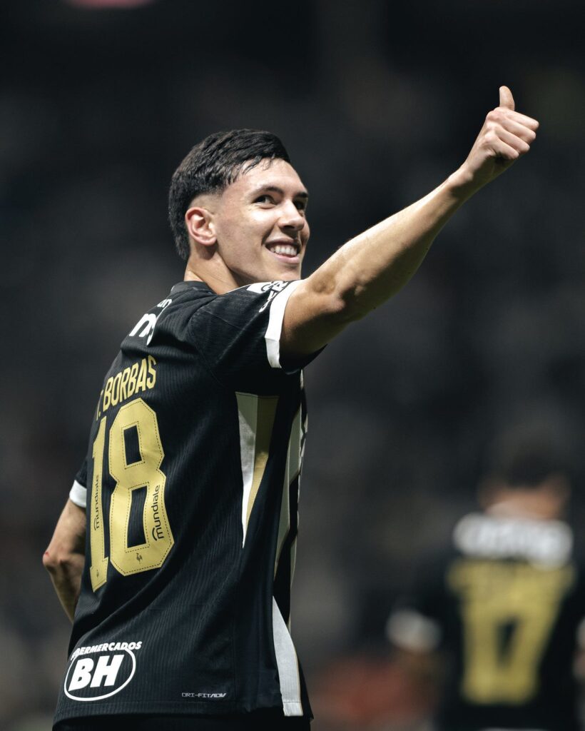Thiago Borbas, de costas, com o nome e o número 18 em dourado na camisa preta, sorri e faz sinal de positivo com o braço erguido
